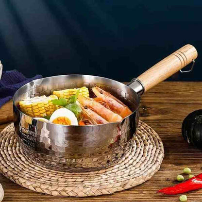 Casserole design en acier inoxydable martelé avec manche en bois, contenant un bouillon de crevettes, maïs et œufs sur un dessous de plat.