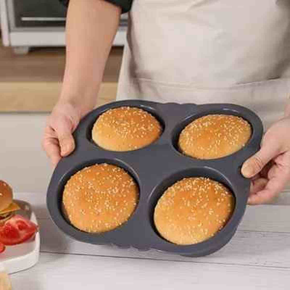 Personne tenant un moule en silicone noir AntiadhésifPro contenant quatre pains burger dorés aux graines de sésame, illustrant la facilité de cuisson.
