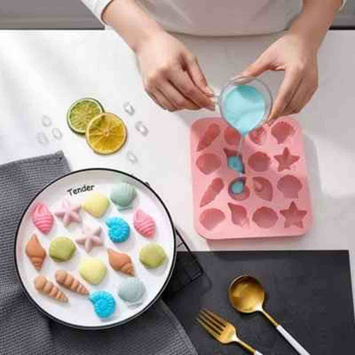 Moule en silicone rose rempli d'une préparation bleue à côté d'une assiette de petits gâteaux en forme de coquillages et étoiles de mer aux couleurs pastel.