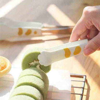 Pince à pâtisserie en forme de patte de chat blanc et jaune saisissant une tranche de gâteau roulé au matcha, idéale pour un service élégant.