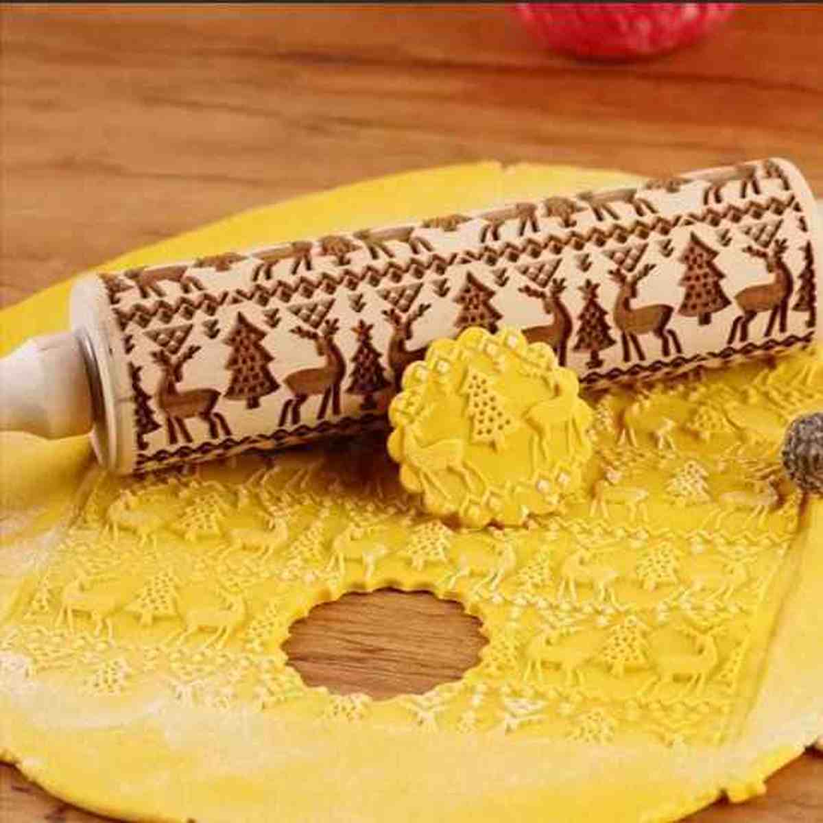 Rouleau à pâtisserie en bois gravé avec des motifs de cerfs et de sapins de Noël, présentant un biscuit en relief assorti sur une pâte étalée.